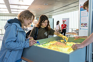 Zwei Menschen stehen an einem Exponat, welches mit Sand gefüllt ist. Sie verschieben mit Kellen den Sand zu Bergen und Tälern. 