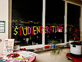 Innenaufnahme eines Flurs bei Dunkelheit mit drei nebeneinanderliegenden Fenstern, auf deren Scheiben buntes Klebeband in Neon­farben die Worte „STUDIENBERATUNG“ bildet. Zwischen den Buchstaben sind kleine weiße Schneeflocken­aufkleber verteilt und im Fenster spiegelt sich die gegenüberliegende, mit Lichterketten geschmückte Wand. Auf der Fensterbank liegen Prospektstapel, rechts auf einem kleinen Tisch ein Heißgetränke­automat. Auf einem Stehtisch links davor liegen Flyer, ein Teller mit Keksen und drei batteriebetriebene LED-Teelichter.