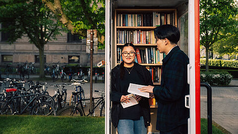 Zwei Studierende stehen am Campus der TU Braunschweig in der geöffneten Tür einer alten telefonzelle, die zum Büchertausch-Schrank umfunktioniert ist. 
