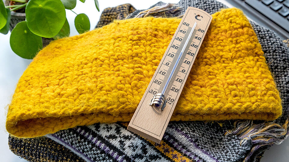 Ein Holzthermometer auf einem Stapel warme Kleidung zeigt 19 Grad Celsius an.