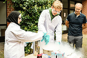 Zwei Studierende in weißen Kitteln mischen Flüssigkeiten in einem Topf zusammen, aus dem es dampft