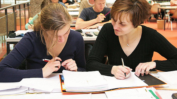 Zwei Studentinnen sitzen zusammen in der Bibliothek und schauen in ein Heft 