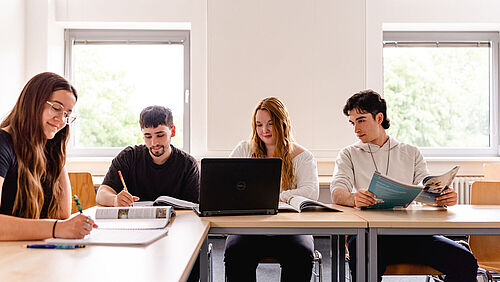 Four students sit in a classroom working on laptops and books.