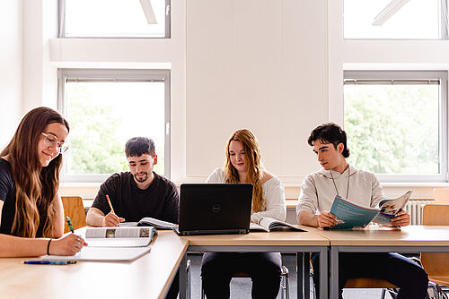 Vier Studierende sitzen in einem Unterrichtsraum und arbeiten an Laptops und Büchern.