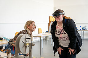Zwei Menschen betrachten ein Gerät, eine Person lacht herzlich und die andere trägt eine Art VR-Brille.