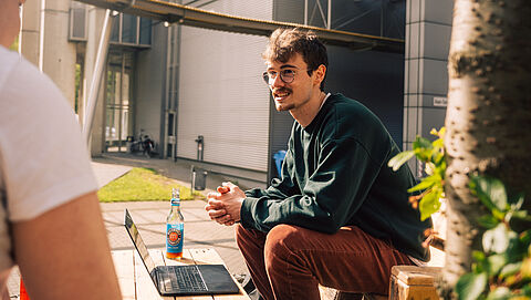 Ein Student sitzt am Campus der TU auf einem Palettensitz im Freien, sein Laptop steht vor ihm auf einem Palettentisch. 