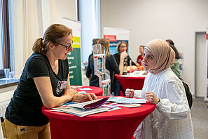 Zwei Menschen stehen an einem roten Stehtisch und unterhalten sich. Auf dem Stehtisch liegen Broschüren. Im Hintergrund sind weitere Gespräche zu sehen