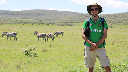 Der Student Nils Michalke steht in einer Steppenlandschaft. Im Hintergrund grasen Zebras.