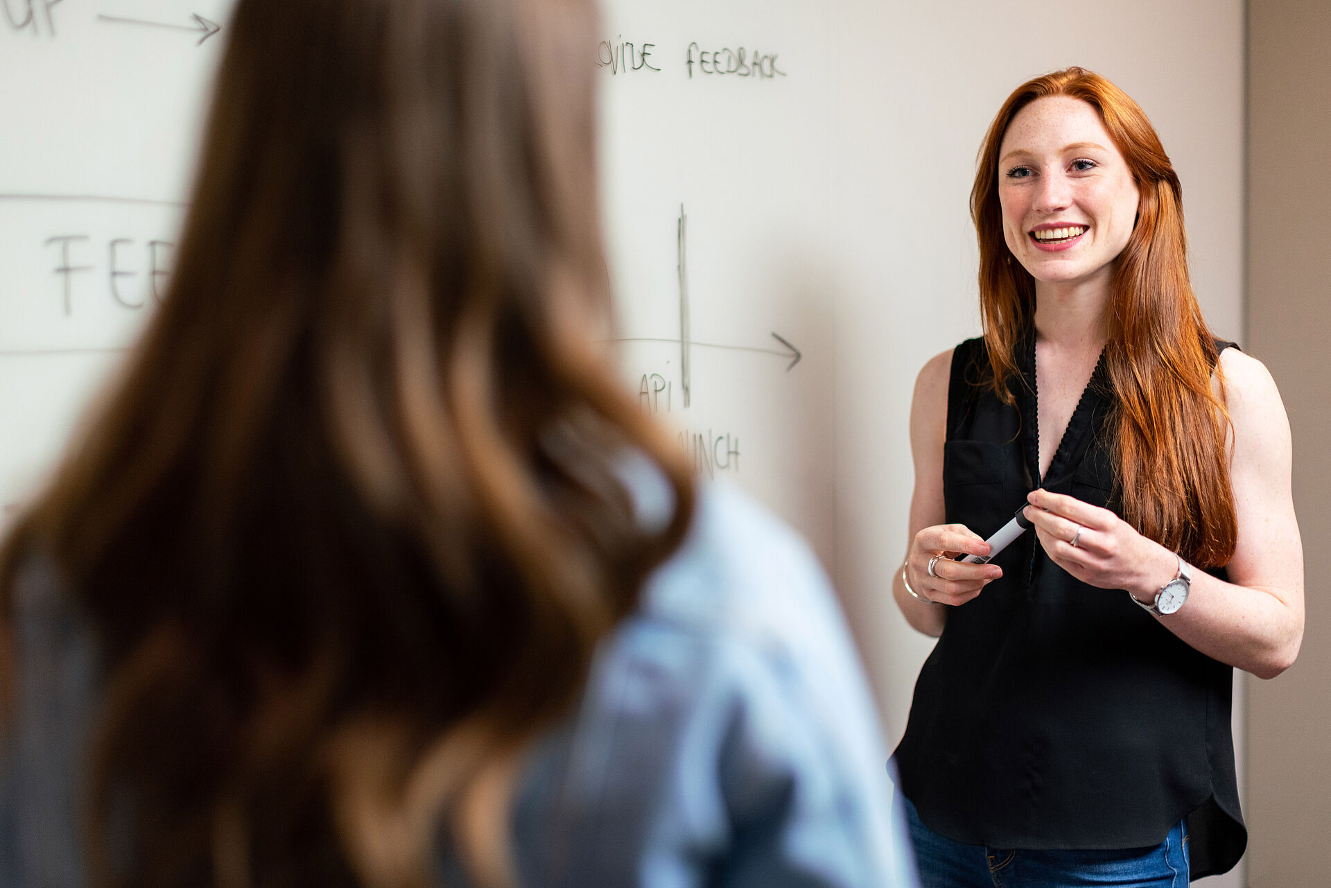 A young woman stands in front of a whiteboard and explains a formula to another woman.