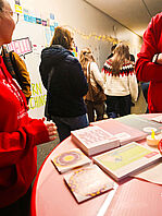 Ein belebter Flur, in dem mehrere Personen mit Winterjacken an einem Info­stand stehen. Im Vordergrund rechts ein Stehtisch mit roter Tischdecke, darauf liegen Broschürenstapel. An der gegenüberliegenden Wand sieht man eine Flurtür, darauf in einer schwarzen Sprechblase der Schriftzug „HELLO“ in Pink. Darunter klebt ein neon-gelber Schriftzug, und über allem verläuft eine warm leuchtende Lichterkette.