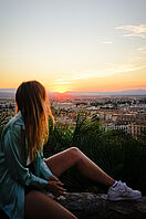 Eine Studentin sitzt auf einem Aussichtspunkt und blickt auf eine spanische Stadt im Sonnenuntergang. 