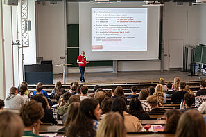 Man sieht einen gut gefüllten Hörsaal, ein Mann in einem TU T-Shirt hält einen Vortrag. Auf der Leinwand sieht man Informationen zur Bewerbung an der TU Braunschweig.