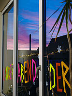 Ein Foto von einem Fenster bei Abenddämmerung: Der Himmel zeigt leuchtende Blau-, Rosa- und Violetttöne. Auf der Scheibe sind aus neonrosa, neongelb und neongrünem Klebeband in Großbuchstaben die Worte „LANGER ABEND DER“ aufgeklebt, ergänzt durch einzelne weiße Schneeflocken-Sticker. 