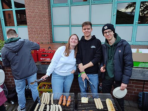 Grillfeier, Drei Studis stehen am Grill und halten Grillzangen