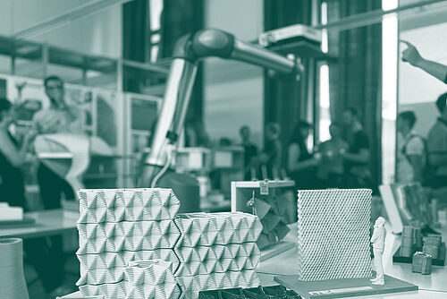 Tisch mit wabenartigen 3D-Druck-Modellen und einem Roboterarm im Vordergrund; im unscharfen Hintergrund unterhalten sich mehrere Personen.