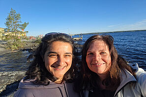 Zwei TU-Mitarbeiterinnen nehmen gemeinsam an einer sonnigen Uferpromenade ein Selfie auf.
