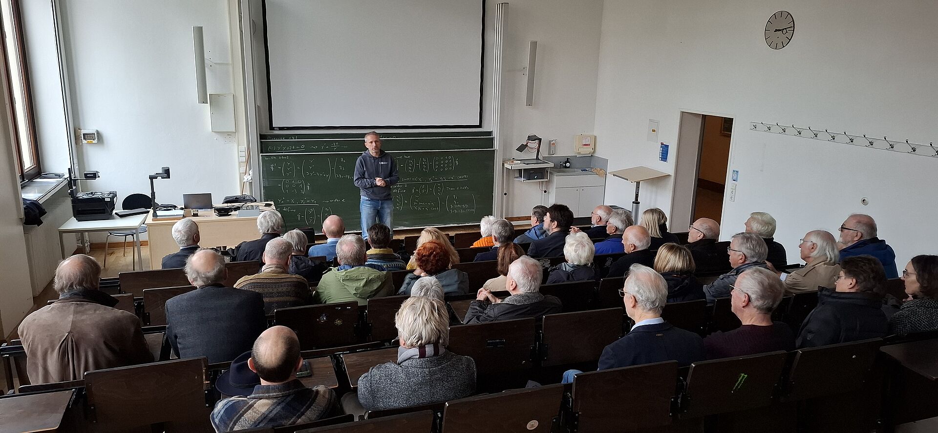 Zu sehen sind Ehemalige des Bauingenieurjahrgangs 1975 in einem Vorlesungsraum mit Herrn Professor Schwerdtner, der präsentiert