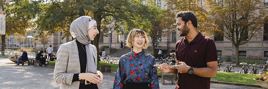 Three students walk across the university square and talk to each other. The old TU Braunschweig building can be seen in the background.