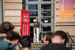 TU Präsidentin Angela Ittel steht auf den Treppen des Altgebäudes. Neben ihr steht ein Roll-Up in TU-rot mit dem Logo der Universität. Am Gebäude hängt eine bunte HIT-Fahne. Frau Ittel ist im Begriff eine Rede zu halten. Im Vordergrund sind viele junge Menschen zu sehen