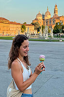 Eine junge Frau isst ein Eis vor einem Park mit großen historischen Gebäuden im Hintergrund.