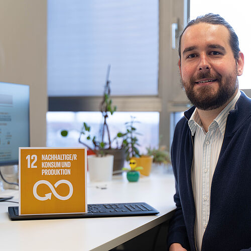 Dr. Christian Weckenborg sitzt an seinem Schreibtisch, auf dem die Karte mit dem 12. SDG zusehen ist.