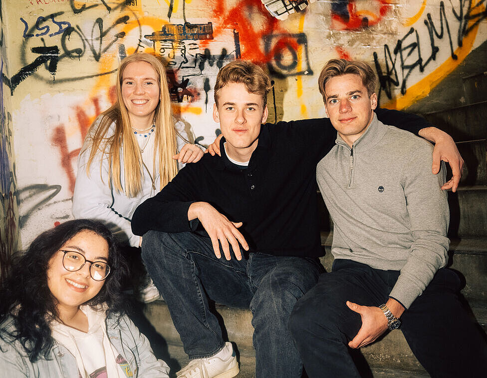 Studierende sitzen auf einer Steintreppe, hinter ihnen eine Wand mit Graffiti