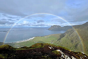 Blick von einer grasbewachsenen Klippe auf das Meer; am leicht bewölkten Himmel spannt sich ein vollständiger Regenbogen über den Horizont.