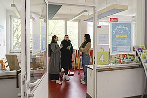 Drei Personen stehen in der Sandkastenzentrale im Altgebäude und unterhalten sich. Im Vordergrund hängt ein Plakat mit der Aufschrift "International Days".