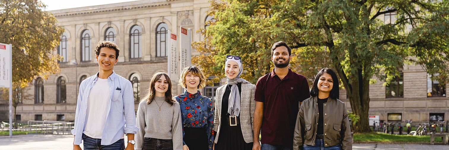 Sechs internationale Studierende gehen über den Universitätsplatz und lächeln in die Kamera. Im Hintergrund ist das Altgebäude zu sehen. 