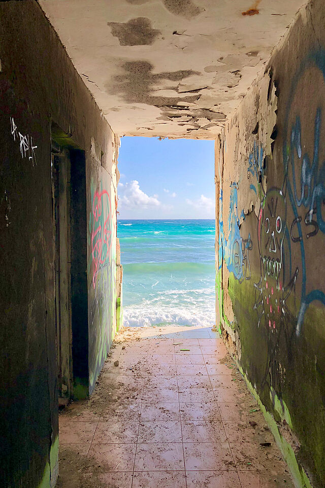 Ein dunkler mit Graffiti beschmierter Gang, an dessen Ende das türkis-blaue Meer vor der Karibikinsel Cozumel zu sehen ist. 