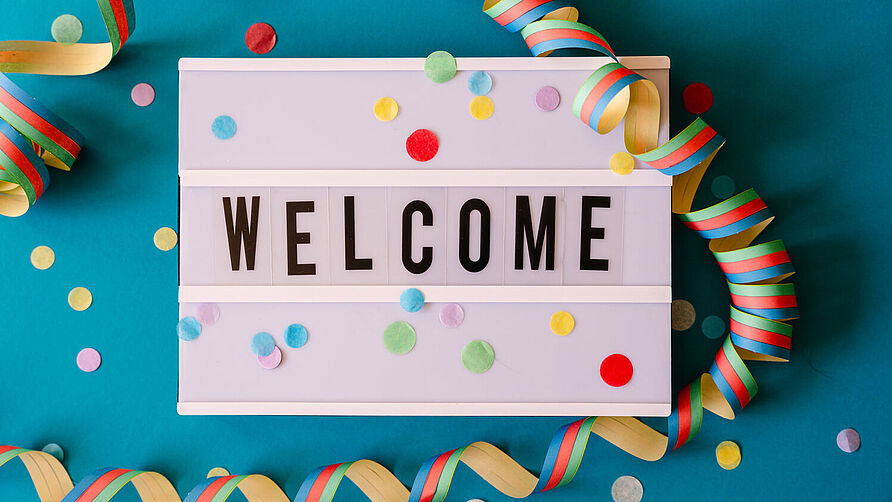Eine Leuchtbox mit dem Schriftzug "Welcome" liegt auf einem blauem Hintergrund, der mit Konfetti und Luftschlangen geschmückt ist.
