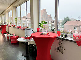 Zu sehen ist ein rot bezogener Stehtisch im Vordergrund, auf dem ein Teller Kekse und ein Aufsteller, der über die Angebote der Zentralen Studienberatung informiert, steht. Dahinter steht ein Punschkochtopf auf einem Tisch und im Hintergrund sind weitere winterlich geschmückte Stehtische zu sehen.