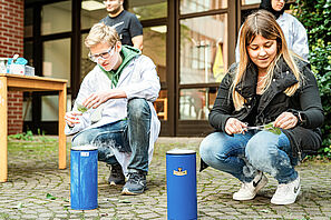 Zwei Menschen hocken vor kleinen Containern aus denen es dampft und halten Blätter in Zangen in den Händen