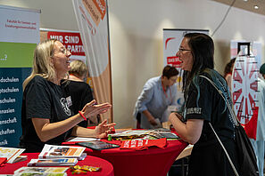 Eine Mitarbeiterin der TU Braunschweig steht an einem Infostand und unterhält sich enthusiastisch mit einer Schülerin. Beide lachen.