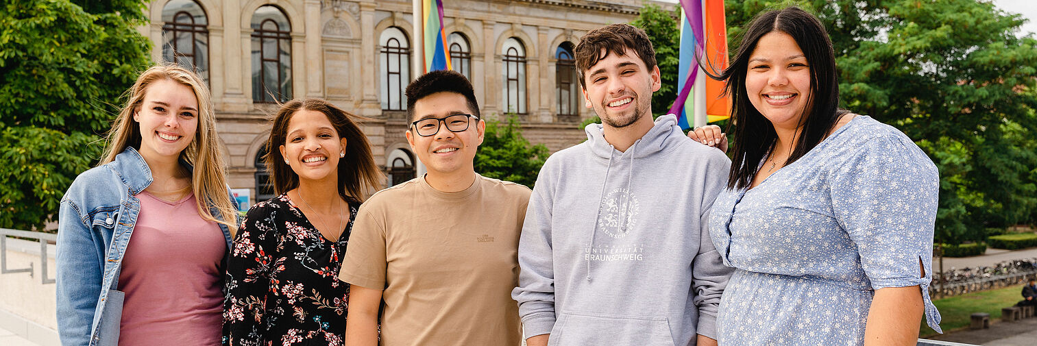 Fünf Studierende stehen nebeneinander auf der Terasse des Audimax. Im Hintergrund ist das Altgebäude der TU zu erkennen.  