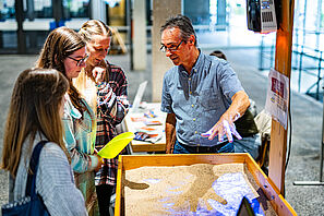 Studieninteressierte stehen an einem Messestand, an dem ein Experiment mit Sand stattfindet