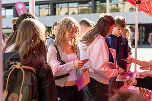 Mehrere junge Menschen stehen an einem Info-Stand. Eine junge Frau mit blonden Locken hält bunte Flyer in der Hand. Andere junde Menschen bekommen weitere Flyer von den Menschen am Info-Stand gereicht.