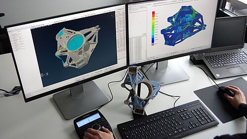 Mitarbeiter sitzt vor zwei Bildschirmen mit CAD-Modellen, vor ihm liegt das fertig gedruckte 3D-Modell.