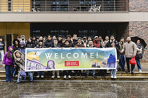 Eine große Gruppe von internationalen Studierenden steht vor dem BRICS und hält ein Banner mit der Aufschrift "Welcome" hoch.