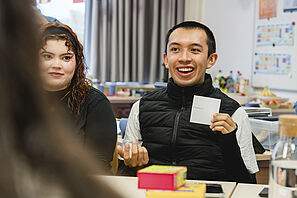  A student laughs cheerfully while sitting at a table with other students and holding up a piece of paper.