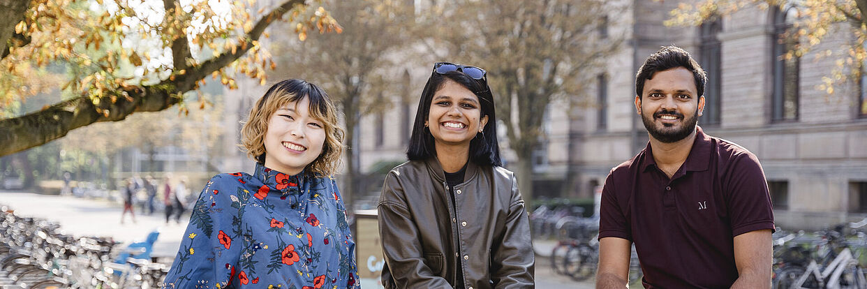 Drei internationale Studierende sitzen vor dem Altgebäude der TU Braunschweig und lachen in die Kamera.  