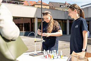 Zwei Personen stehen auf dem Universitätsplatz. Vor Ihnen ist ein Tisch, auf dem ein Experiment mit vielen kleinen Ampullen aufgebaut ist