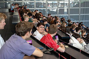 Schüler und Schülerinnen sitzen in einem Hörsaal und lauschen einer Vorlesung. Ein Schüler schaut freundlich lächelnd in die Kamera.