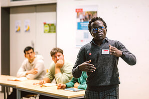A speaker talks with commitment during a workshop. Two workshop participants are sitting in the background.