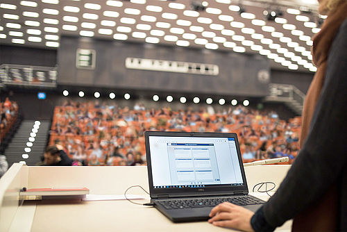 Laptop im Vordergrund; im Hintergrund sitzen Studierende im Audimax
