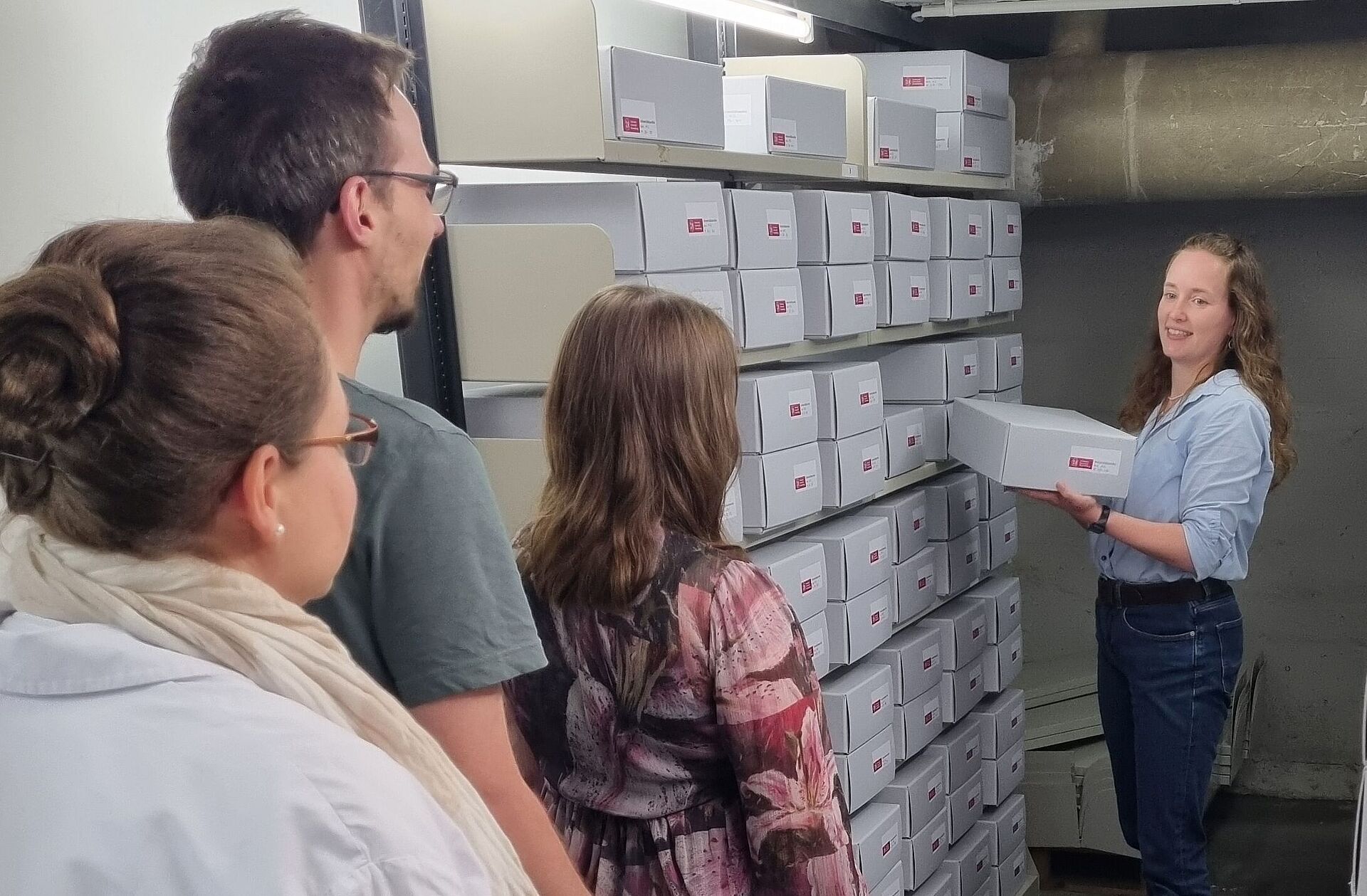 Zu sehen ist die Archivleitung, Tanja Wolf, vor einem Archiv-Regal, zu drei anderen Menschen (in Rückenansicht) sprechend.