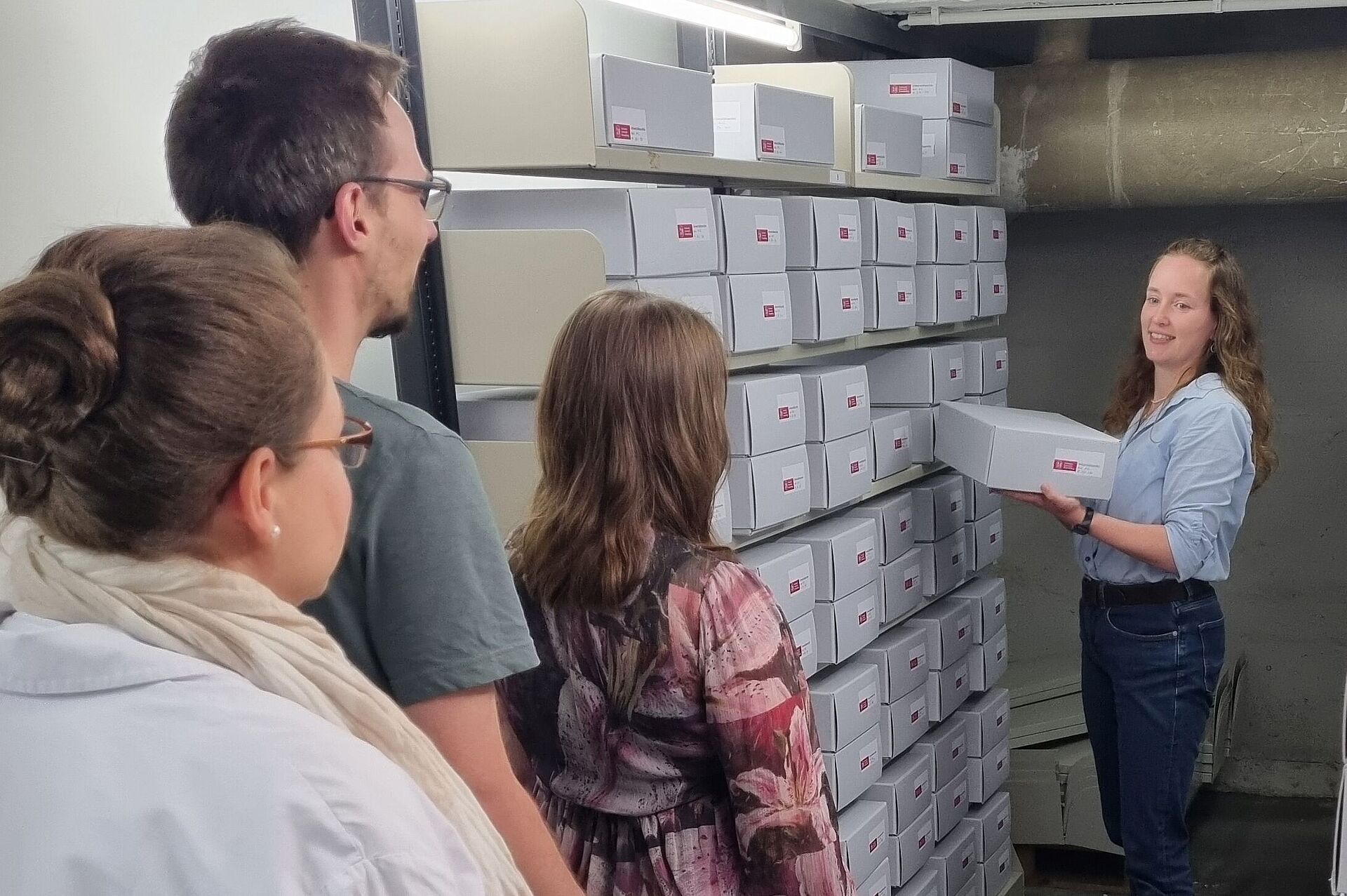 Zu sehen ist die Archivleitung, Tanja Wolf, vor einem Archiv-Regal, zu drei anderen Menschen (in Rückenansicht) sprechend.