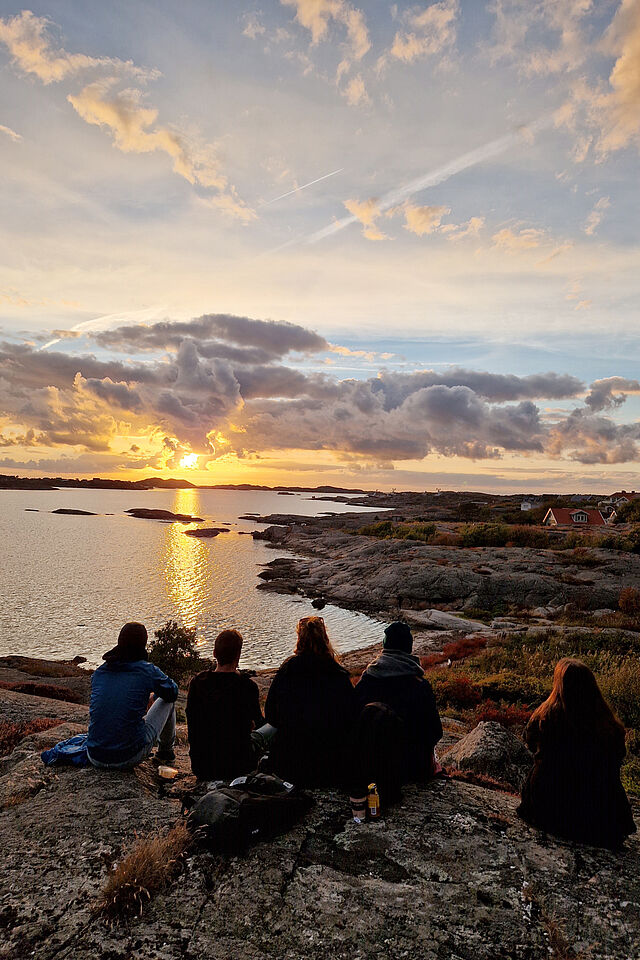 A group of students sits on a hill and watches the setting sun. 