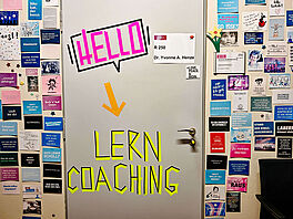 Auf einer grauen Tür klebt ein schwarzer Umriss einer Sprechblase mit dem neon-pinken Schriftzug HELLO, darunter ein breiter gelber Pfeil nach unten zu den in leuchtend gelbem Klebeband geformten Buchstaben LERN COACHING. Links und rechts der Tür sind die Wände mit dicht an dicht angeordneten Postkarten beklebt, auf denen kurze Sprüche und Stichworte stehen.