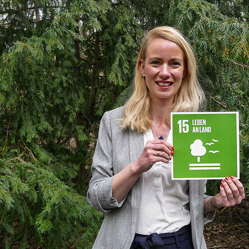 Dr. Ruthsatz steht im Grünen und hält das Schild für das 15. SDG. 